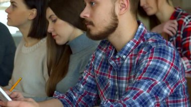 Sakallı bir erkek öğrenci yüzünü kameraya doğru çeviriyor, canlı ve kareli bir gömleğe güvenerek. Etrafı odaklanmış bayan sınıf arkadaşlarıyla çevrili bu yakışıklı lisans öğrencisi modern öğrenmenin özünü canlı bir konferans salonunda yakalıyor.