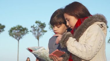 İki meraklı çocuk, bir oğlan ve bir kız, açık mavi bir gökyüzünün altında dışarıda dururken haritayı dikkatle inceliyorlar. Sıcak, beyaz kürklü bir ceket ve canlı kırmızı bir eşarp giyen kızla birlikte bir sonraki maceralarını ararken sıcacık giyinmişler.