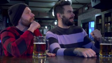 İki sakallı arkadaş, biri kareli gömlekli, diğeri çizgili kazaklı, bardaki favori spor takımlarına tutkuyla tezahürat yapıyorlar. Önlerinde soğuk bardaklar dolusu birayla, heyecan içinde yumruklarını kaldırıyorlar, heyecanı somutlaştırıyorlar.