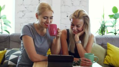 Yeşilliklerle dolu rahat bir kafede iki neşeli kız tabletlerindeki bir şeyden büyülenip kahkahalarını ve neşelerini paylaşıyorlar. Rahatlatıcı tavırları ve parlak gülümsemeleri harika bir kahve molası için mükemmel bir ortam yaratıyor.