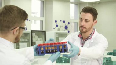 Beyaz önlüklü ve lateks eldivenli iki genç erkek laboratuvar teknisyeni, bilimsel araştırmalarda takım çalışması sergileyerek birlikte test tüpleri alışverişinde bulunuyorlar. Parlak, steril ortam gelişmiş ekipmanlarla dolu.