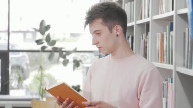 Büyüleyici genç bir adam parlak bir kütüphanede duruyor, parlak turuncu bir kitaba dalmış, düşüncelere dalmış. Bilgi raflarıyla dolu huzurlu atmosfer yaratıcılığa ve hayal gücüne ilham verir.