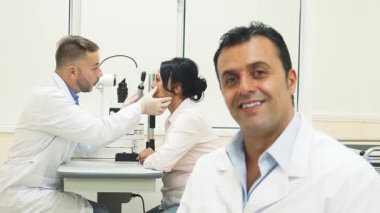 Neşeli bir doktor güven içinde ön planda oturur, sıcaklığı ve yakınlığı yayar. Arkasında, odaklanmış bir göz doktoru hastaların gözlerini özenle inceliyor, modern bir tıp ofisinde profesyonel ve ilgili bir atmosfer sergiliyor.