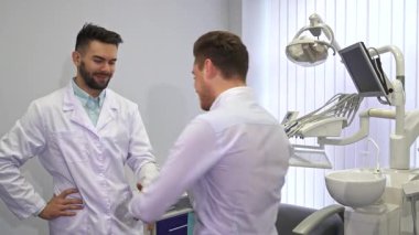 Yakışıklı sarışın bir hasta modern bir klinikte genç bir erkek dişçiyle diş sorunlarını tartışırken hareketli bir şekilde hareket eder. Diş hekimi dikkatlice dinler ve gelişmiş diş hekimleriyle çevrili sıcak bir güven ve bakım ortamı yaratır.