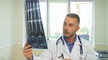 Beyaz laboratuvar önlüklü odaklanmış genç bir adam X-Ray görüntüsünü havada tutuyor ve iyi aydınlatılmış ofisinde inceliyor. Boynuna sarılı steteskop profesyonel atmosfere katkı sağlarken, bej duvarlar ve modern mobilyalar...