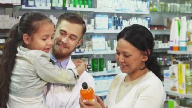 Parlak bir eczanede, dost canlısı bir eczacı, neşeli küçük bir kızı annesi canlı turuncu bir ilaç tüpünü incelerken ziyaret eder. Sıcak etkileşim, sağlık ve güzellik seçeneklerini birlikte incelerken aile bağlarını vurguluyor.