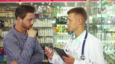 Beyaz önlüklü, odaklanmış bir doktor elinde bir tablet ve haplarla enerjik bir eczanede bir hastayla düşünceli bir tartışmaya girişiyor. Hasta, ilginç bir ifadeyle, dikkatle dinliyor, sağlık ve güven dinamik bir atmosfer yaratıyor.