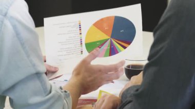 Kesilmiş bir dikiz aynası, modern ofis ortamında kahvenin tadını çıkarırken canlı basılı bir diyagramı inceleyen iki profesyonel adamı derinden tartışırken yakalıyor. Düşünceli ifadeleri strateji kurarken işbirlikçi bir ruhu yansıtıyor.