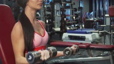 Uzun sarı saçları parlak at kuyruğu olan güçlü kız, iki ağır dambılı rahatça kaldırır ve forma olan bağlılığını gösterir. Odaklanmış ifadesi ve atletik kıyafeti, enerjik bir spor salonunda güç eğitimine olan bağlılığını vurguluyor.