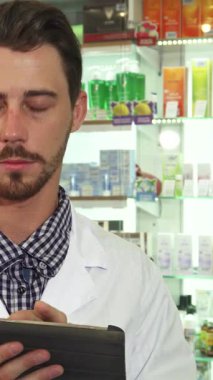 Gıcır gıcır beyaz bir tıbbi elbise giymiş odaklanmış bir erkek eczacı, renkli ilaçlarla dolu canlı raflarla çevrili şık tabletindeki ilaç envanterini özenle inceliyor. Onun yoğun bakışları ve profesyonel tavırları,