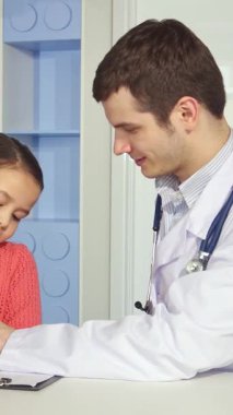 Neşeli bir Kafkas erkek çocuk doktoru parlak gözlü küçük bir kızla samimi bir şekilde yakınlaşır, canlı bir pediatri ofisinde bir panoya resim çizer. Renkli çevre ve bulaşıcı gülümseme neşe ve güven dolu bir atmosfer yaratır.