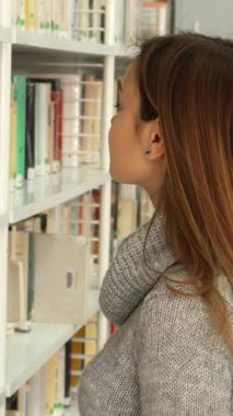 Uzun kahverengi saçlı, rahat gri bir kazak giyen odaklanmış beyaz bir kız kütüphanedeki düzenli raflar arasında bir kitap aramakla son derece meşgul. Sahne, rengarenk yerlerde dolaşırken konsantrasyonunu yakalıyor.