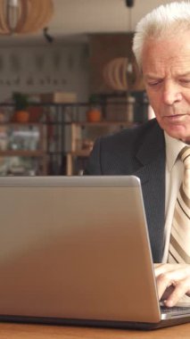 Özel dikim takım elbiseli ve çizgili kravatlı zarif, yaşlı beyaz bir adam laptopuna dalmış bir şekilde kırsal ahşap bir masada oturuyor ve dumanı tüten bir fincan kahvenin tadını çıkarıyor. Kafenin davetkar atmosferi, yumuşak ışıklandırma ve şıklıkla bezenmiş.