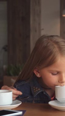 Sıcak ve davetkar bir kafede, sevimli küçük bir kız çayının tadını narin beyaz bir fincandan çıkarırken, annesi büyüleyici bir hikayeyi paylaşıyor. Yumuşak ışıklandırma ve rahat dekor, neşeli yakınlaşma anları için mükemmel, samimi bir atmosfer yaratır.