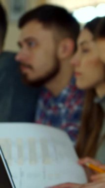 Büyüleyici bir kız öğrenci, kendini derse daldırırken koyu renk saçlarıyla yüzünü çerçeveliyor. Bu yakın plan, onun canlı akademik ortamda sınıf arkadaşları tarafından çevrelenmiş konsantrasyon ve adanmışlığını vurguluyor.