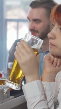 Kızıl saçlı genç bir kadın, arkadaşları yakınlardaki animasyon sohbetleriyle meşgulken hareketli bir barda tek başına oturmuş, buzlu bir bardak bira içiyor. Hüzünlü ifadesi, hayat dolu gece hayatının ortasında dokunaklı bir yalnızlık hissi yakalıyor.