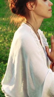 Huzurlu genç bir kadın zarif bir yoga pozunda oturur, ellerini meditasyonda birbirine bastırır, huzur ve farkındalığı somutlaştırır. Canlı yeşil çimlerle çevrili ve ılık yaz güneşiyle yıkanmış, sakin bir enerji yayıyor, sükuneti buna davet ediyor.