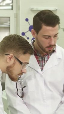 Gösterişli, iyi ışıklandırılmış bir laboratuvarda, gevrek beyaz önlüklü ve mavi lateks eldivenli iki genç erkek asistan iş akışları hakkında animasyon bir tartışmaya giriştiler. Odaklanmış ifadeleri ve çevrelerindeki parıldayan bilimsel ekipmanları