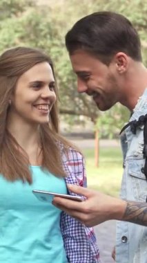 Neşeli genç bir kadın ve yakışıklı dövmeli bir adam, güneşli kampüs yolunda yan yana dururken, akıllı telefonlarından büyülenmiş bir şekilde keyifli bir anı paylaşıyorlar. Işıl ışıl gülümsemeleri ve çevrelerindeki canlı yeşillik bir atmosfer yaratıyor.