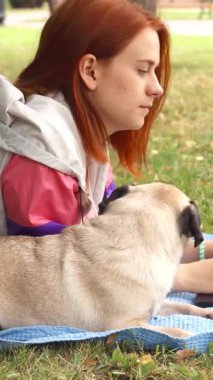 Çarpıcı kızıl saçlı genç bir kız, güneşli bir parkta canlı mavi bir battaniyenin üzerinde dizüstü bilgisayarında dikkatle yazı yazıyor. Onun yanında, büyüleyici köpeği dünyayı meraklı gözlerle izliyor, keyifli bir arkadaşlık ve eğlence anını somutlaştırıyor.