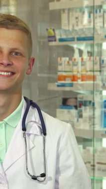 Neşeli bir erkek doktor, kendine güvenen bir şekilde parlak bir eczanede duruyor ve elinde bir paket tutarken ışıldıyor. Yumuşak beyaz önlük ve steteskop ile tamamlanmış yaklaşımına bakılırsa renkli bir arkaplan karşısında sıcaklık ve profesyonellik yayıyor.