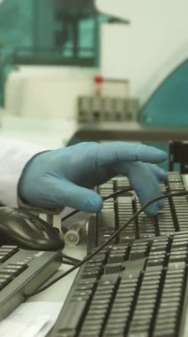Kendini işine adamış beyaz önlüklü ve mavi lateks eldivenli bir erkek laboratuvar teknisyeni test tüplerinden düzgün bir bilgisayara ustalıkla veri giriyor. Bu canlı sahne teknoloji ve biyolojinin kesişimini vurguluyor, hassasiyeti vurguluyor ve