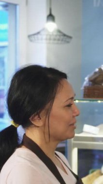 Enfes bir fırın ortamında, olgun bir Asyalı fırıncı kadın sıcak bir somun ekmeği yakışıklı erkek müşterisine uzatarak gerçek bir bağ kuruyor. Artistik ekmek ve yumuşak aromalı davetkar atmosfer.