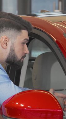 Şık, sakallı bir adam galeride göz kamaştırıcı kırmızı bir arabayı incelerken neşe saçıyor. Çekici gülümsemesi yeni otomotiv hazineleri keşfetmenin heyecanını yakalıyor. Zekice giyinerek modern tüketiciliğin heyecanını somutlaştırıyor.