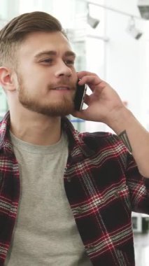 Son moda ekose gömlekli neşeli, sakallı bir adam şık bir araba galerisinde akıllı telefonuyla konuşuyor. Kendine güvenen gülüşü ve parlak gözleri araba alışverişinin heyecanını yansıtıyor. Parlak araçlarla çevrili.