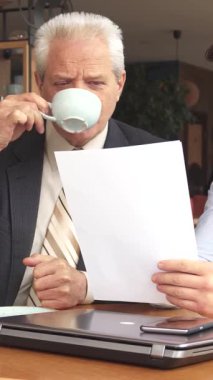 Sıcak ve davetkar bir kafede, iki erkek iş ortağı, önemli belgeleri analiz etmek için odaklanmış bir tartışmaya daldılar. Beyaz saçlı kıdemli beyaz işadamı, çayını yudumlarken genç adam keskin bir takım elbise giymişti.