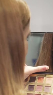 Aynadan yansımasına ışıldayan genç bir kadının yakın plan fotoğrafı. Makyajını elinde paletle ustalıkla yapıyor. Bu canlı sahne kendini ifade etmenin ve güzelliğin neşesini özetliyor. Kusursuz cildini ve şık saçlarını gözler önüne seriyor.