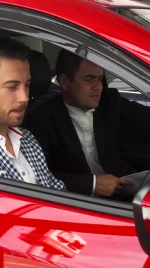 Çarpıcı kırmızı bir arabanın içinde, şık giyimli bir satıcı ve özenli bir alıcı modern iç mekanı eşeliyor, yüksek teknoloji gösterge panelini ve kontrolleri inceliyor. Canlı gösteri salonu atmosferi, lüks arabalar hakkındaki ilgi çekici tartışmalarını güçlendiriyor.