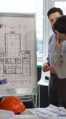 Karizmatik genç bir erkek mimar ayrıntılı bir bina planına hevesle el kol çizelgesi üzerinde yaklaşarak meslektaşlarını güneşli bir ofiste büyülüyor. İki odaklı inşaat mühendisi, biri şapka takıyor, her iki tarafta duruyor, derinden.
