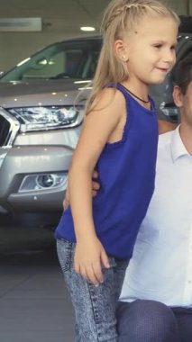 Mutlu bir baba, parlak bir galeride göz kamaştırıcı kırmızı bir arabayı keşfederken her ikisi de heyecan saçan kızının yanında diz çöker. Neşeli etkileşimleri, kahkahalarla ve sevgi dolu jestleriyle, iç açıcı bir aile anını yakalar.