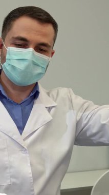 Maske ve eldiven takmış, kendini adamış esmer, beyaz dişçi titizlikle orta yaşlı bir kadın hastanın detaylı muayenesine hazırlanıyor. Yakın plan, dişçilerin elinde bir orak probu tutarken odaklanmış ifadesini yakalar.