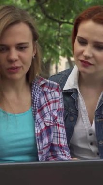 Canlı üç genç öğrenci, iki kadın ve bir erkek, verimli yeşilliklerle çevrili, güneşli bir kampüs bankında dizüstü bilgisayarla neşeyle meşguller. Işıldayan gülümsemeleri ve hareketli ifadeleri dostluk ve işbirliği için davetkar bir ortam yaratıyor.