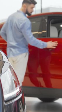 Açık mavi gömlekli şık sakallı bir adam, çağdaş bir otomobil bayisinde çarpan kırmızı bir arabayı dikkatle inceliyor. Görüntü heyecan ve entelektüellik yayıyor, ince çizgileri ve aracın parlak görünümünü yansıtıyor,