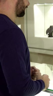 Zarif bir mücevher mağazasında, ışıl ışıl gülümseyen bir bayan satıcı güzel bir şekilde paketlenmiş bir alışverişi zevkle elinde şık bir alışveriş çantası tutan genç sakallı bir adama teslim eder. Arkaplanda pırıl pırıl pırıl mücevherler.