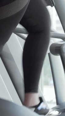Şık, forma uygun spor kıyafetleri içinde canlı, iri yarı bir kadın eliptik bir eğitmen üzerinde enerjik bir şekilde çalışıyor, pozitiflik ve kararlılık yayıyor. Yakın çekim onun neşeli ifadesini ve bacaklarının dinamik hareketini vurguluyor.