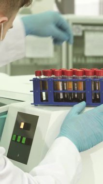 Kendini işine adamış beyaz önlüklü ve parlak mavi lateks eldivenli bir erkek laboratuvar teknisyeni son model bir santrifüje yerleştirmeden önce renkli test tüplerini dikkatle inceliyor. Bu dinamik sahne kesinliğin özünü yakalar.