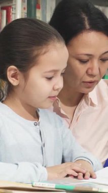 Özenli bir Asyalı anne, sıcak, davetkar bir kütüphanede, hayat dolu kitapların arasında ödevlerini yapmasına yardım ederek tatlı kızına yakın duruyor. Bu dokunaklı an, sevgi ve öğrenme bağını güzel bir şekilde gösteriyor.