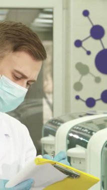 Çıtır beyaz önlüklü ve mavi lateks eldivenli genç erkek laboratuvar asistanı, test sonuçlarını canlı sarı bir klasörde özenle belgeliyor. Son teknoloji laboratuvar ekipmanlarıyla çevrelenmiş, hassasiyet ve adanmışlığı somutlaştırıyor