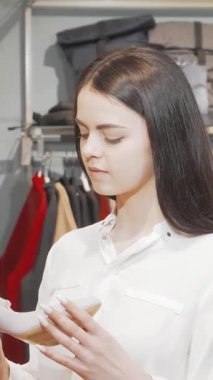 Uzun siyah saçlı zarif bir kadın modaya uygun bir butikte şık yüksek topuklu ayakkabıya dikkatle bakıyor. Canlı giysi ve aksesuarlarla çevrelenmiş, modern zarafeti ve kişisel tarzını özetliyor, düşünceli bir karar vererek