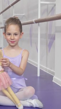 Bu büyüleyici uzunluktaki çekim neşeyle el sallayan küçük bir balerinin etek giymiş bebeğini sallamasını içeriyor. Bir bale stüdyosunun canlı arka planına bakan leylak rengi taytlı ve yumuşak taytlı bu güzel kız...