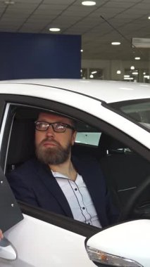 El değmemiş beyaz bir hatchback 'in içinde, keskin siyah takım elbiseli şık bir satış müdürü, sözleşmenin detaylarını tutkulu bir şekilde odaklanmış bir müşteriye açıklar. Sakallı adam, gözlük takıyor, dikkatle dinliyor, profesyonellik ve güven atmosferini teşvik ediyor.