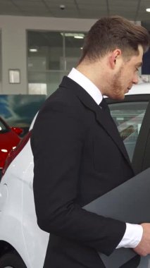 Galerideki el değmemiş beyaz bir hatchback 'in içinde, keskin siyah takım elbiseli şık bir satış müdürü, sözleşmenin detaylarını odaklanmış bir müşteriye tutkuyla anlatıyor. Atmosfer profesyonellik ve güvenle dolu, çekici sakallı adam olarak.