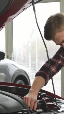 Şık ve bakımlı sakallı genç bir adam lüks bir arabanın parıldayan kaputunun üzerine eğiliyor, güçlü motoru tatmin edici bir gülümsemeyle inceliyor. Doğal ışıkla dolu çağdaş bayilik arka planı, arabanın heyecanını arttırıyor.