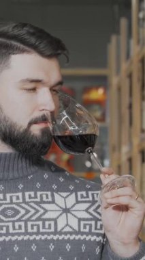 Şık sakallı bir adam bir bardak koyu kırmızı şaraptan zevk alır, anlayışlı bakışları hareketli bir şarap süpermarketinin canlı atmosferini yansıtır. Etrafında zarif şişeler, rahat süveteri ve kendine güveni gerçek bir şarabı somutlaştırıyor.