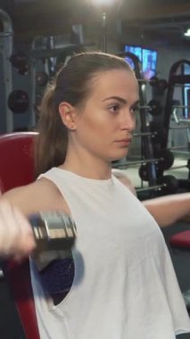 Kararlı genç kadın, uzun sarı saçları geriye doğru kıvırcık at kuyruğu ile, yoğun bir şekilde dambılları kaldırıyor, etkileyici pazılarını ve sarsılmaz bağlılığını sergiliyor. Işıldayan spor aletleriyle çevrili, fitness ruhunu somutlaştırıyor.