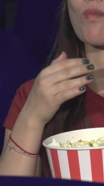 Uzun siyah saçları ve parlak bir gülümsemesi olan keyifli genç bir kadın, klasik kırmızı ve beyaz çizgili kovadan patlamış mısırın tadını çıkarırken sinematik deneyime daldı. Onun günlük kırmızı gömleği tiyatronun canlı atmosferini tamamlıyor.